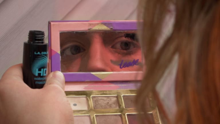 From behind, a person looks into the mirror of an eyeshadow palette and both their eyes are reflected in the mirror. They are applying mascara