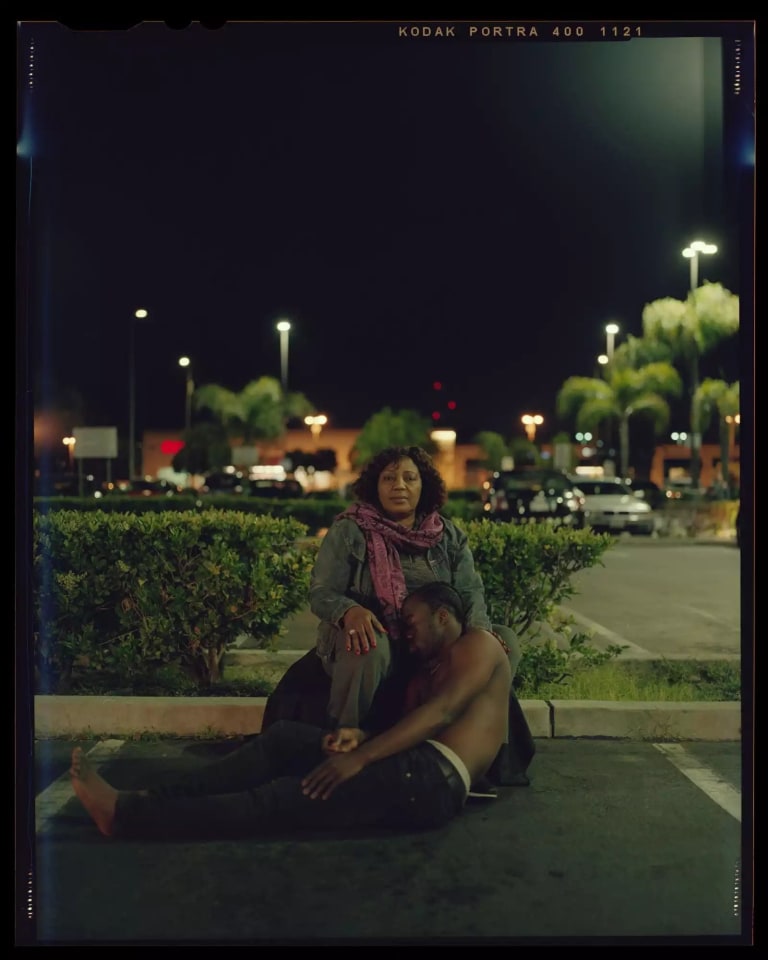 A color photograph of a black woman sitting on the curb in a parking lot, with a shirtless black man resting against her knee and lap. In the background there is a shopping plaza, cars, streetlamps, and palm trees.