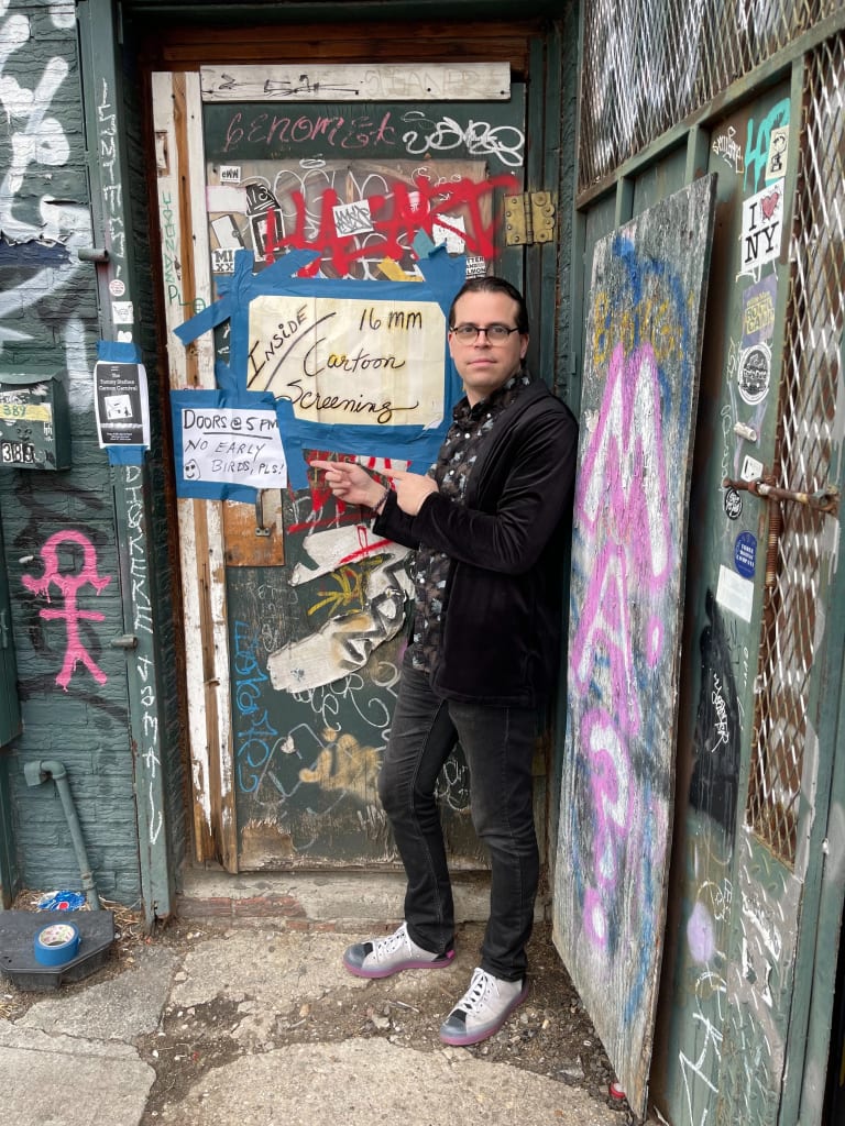 Picture of a man standing in front of a door covered in posters and graffiti, pointing to a poster that reads "Inside: 16mm Cartoon screening. Doors at 5pm, no early birds."