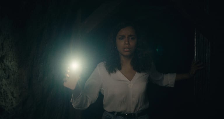 A film still of a terrified-looking young woman making her way through a dark tunnel, using the flashlight on her phone to guide her way.