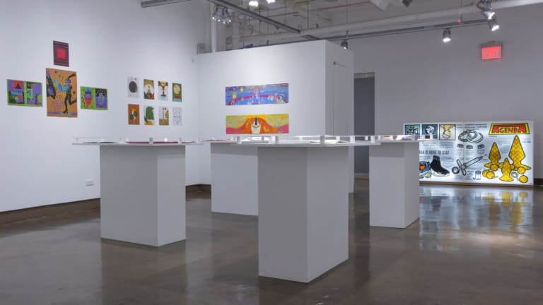 Photo of printmaking artwork hung from inset walls, with pedestals in middle of room displaying books and a large lightbox print on plexiglass to the far right.