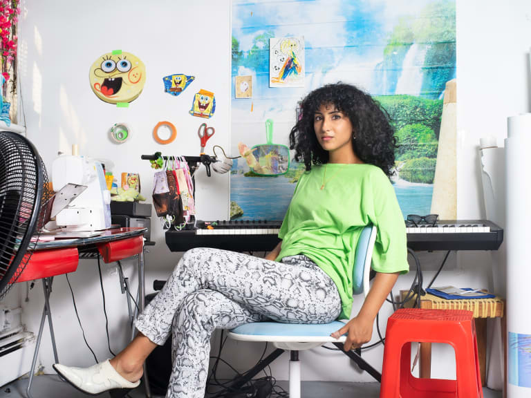 A photograph of Farah Al Qasimi. Farah is sitting on a chair in her studio. In the background are a piano keyboard, sewing machine, and other miscellaneous objects and artwork.
