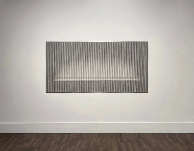 Photo of a white wall in an art gallery, on which is hung a black and white drawing in charcoal and graphite on a sheet of paper. The drawing fills the entire sheet, and consists of very closely spaced vertical lines in varying shades that create the illusion of an alcove receding back into space.