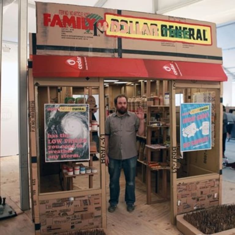 A man standing in a cardboard rendition of popular dollar stores. He is standing in the middle of the "doorway" wearing a button-up shirt and blue jeans.