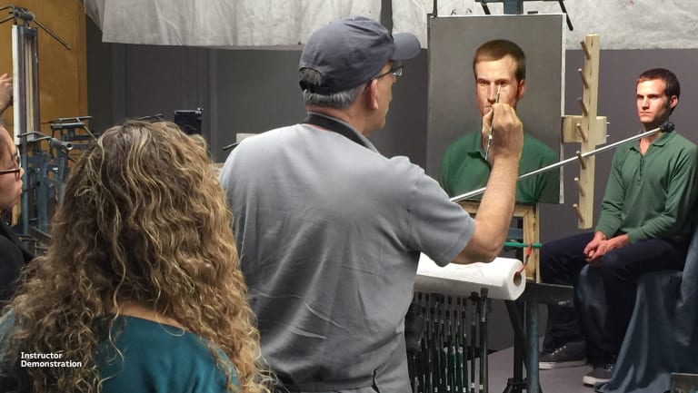 A photograph of a fine arts teacher demonstrating how to paint the portrait of the man posing in front of them to a student standing behind him.