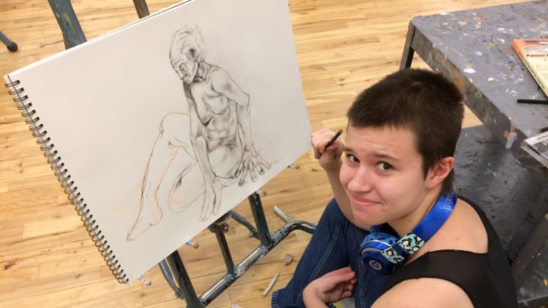 A student pauses drawing a figure to smile at the camera 