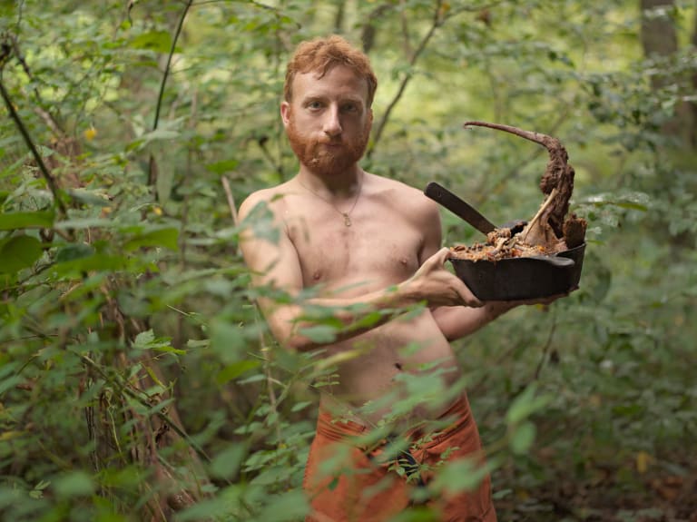 Color photograph of shirtless man surrounded by green foliage in a forest. He is wearing worn orange pants and has red hair and a red beard. He’s holding a deep cast-iron skillet with stew and a large bone in it. 