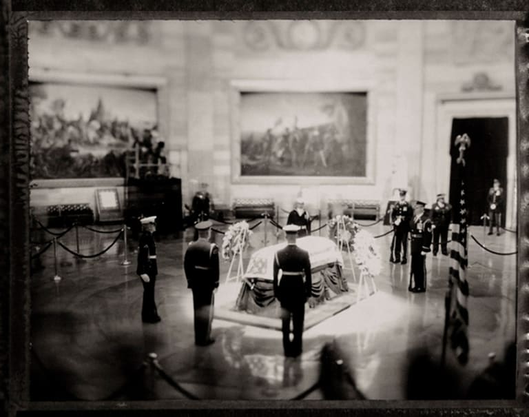 A black and white photo of a casket surrounded by men in military uniform. They are in a large, marble room of the Capitol building. The casket is spotlit and there are paintings on the walls surrounding it.