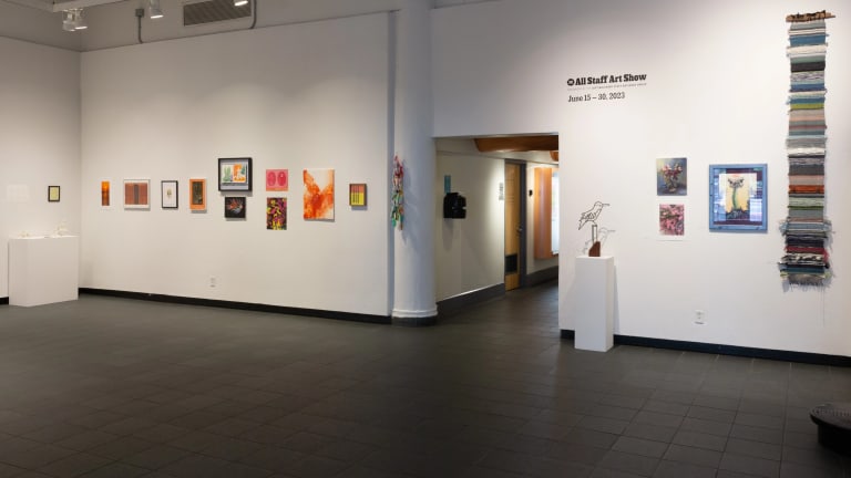 Artwork hung in a horizontal row across a long gallery wall, with a doorway leading to offices in the center of it.