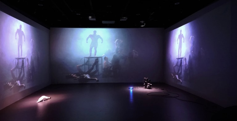 Picture of an installation with three walls being projected with the same image: A foggy dimly lit environment, with several people strewn about the floor, and a single figure of a strong man standing, backlit, against the only source of light.