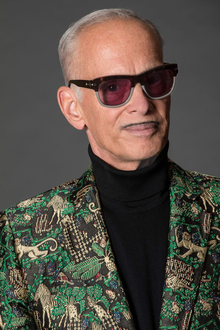 A photograph of John Waters wearing a green suit and red glasses