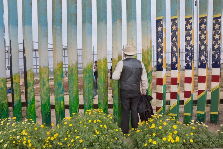 Color photograph of a man in slacks, shirt, vest and a hat looking through the slats of the U.S.-Mexico border fence at people on the other side. The metal slats of the fence are painted with plant motifs and the US flag. 
