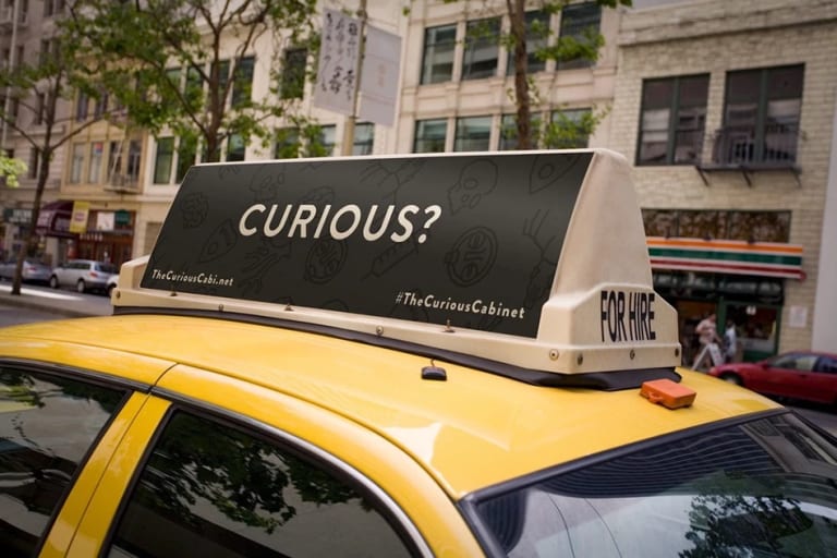 A closeup of a taxi top ad that says "Curious?" in white all caps text on a solid black background