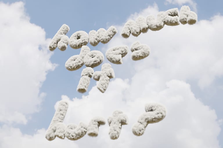 A computer-rendered image of the phrase How Much Does It Hurt? in cloud-like letters hovering before a slightly-cloudy blue sky