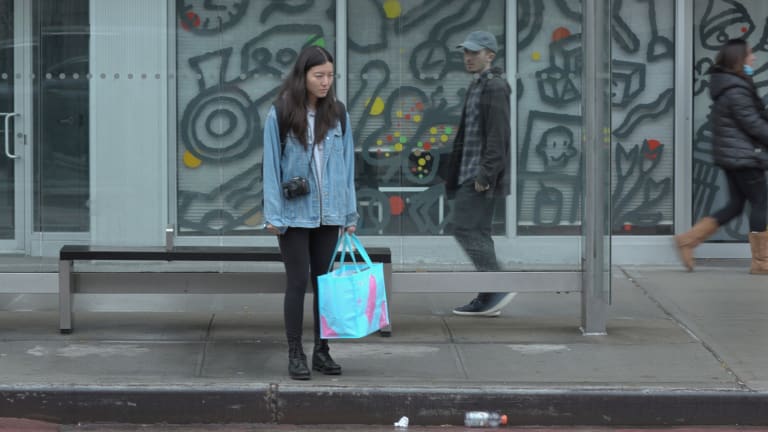 East Asian woman wearing denim jacket stares at the ground waiting for the bus as a white man in the background glares.