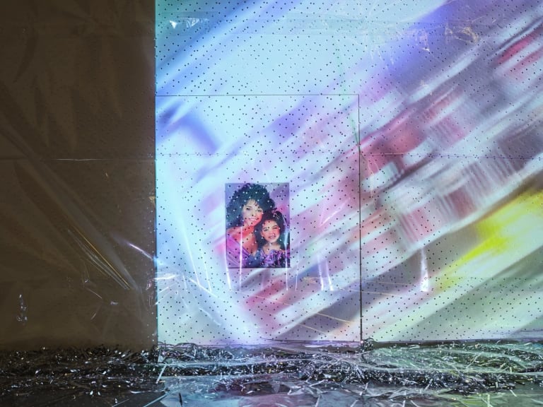 a painting of a woman and young girl in a white installation space, shrouded by cellophane and projected colorful images 