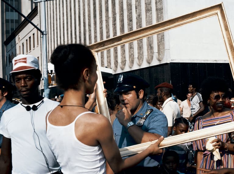 A crowd of black people and a white police officer stand on a street. A woman with her back to the viewer holds up an empty picture frame