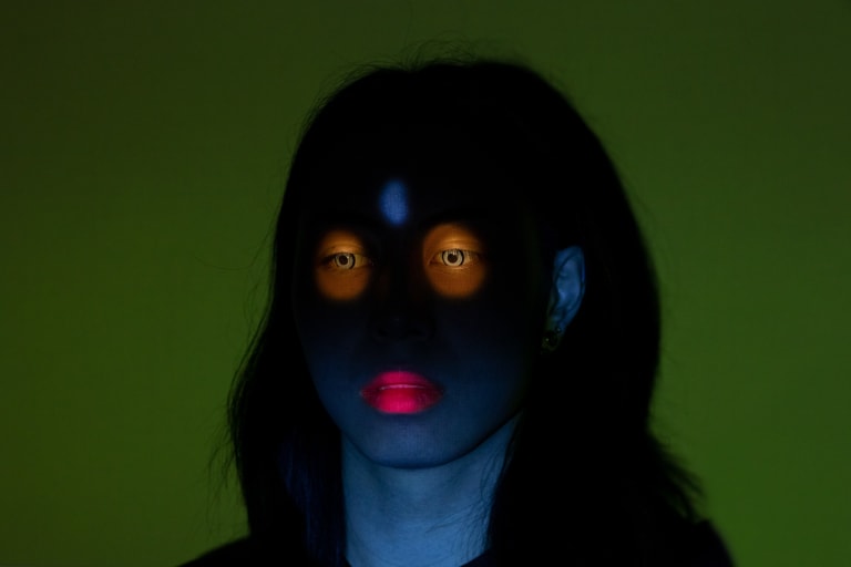 A person from the neck up has a very shaded-out face; the parts of their skin that shows is blue, their long hair is black, their lips are pink, and their eyes are surrounded by an orange glow.