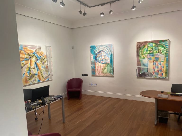 Installation view of three paintings in a corner of an art gallery with white walls. There is a brown chair in the corner, a metal table with a computer on it to the left, and a wooden desk to the right. The desk and table are only half visible, as they are cropped by the sides of the photo.