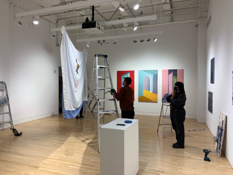 Two people in masks and dark clothes stand in the middle of a gallery space cluttered with latters, pedestals, and artowrks that are bging installed in the space