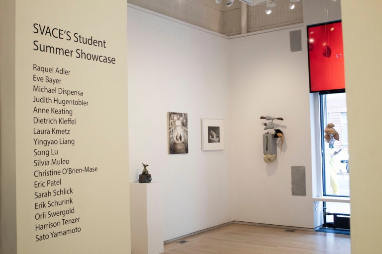 View of the entryway at the “SVACE Student Summer Showcase” at the Flatiron Project Space, 2023. There is a list of names on the wall of the participants in the show. Three sculptural pieces are seen, and two artworks framed on the wall are seen.