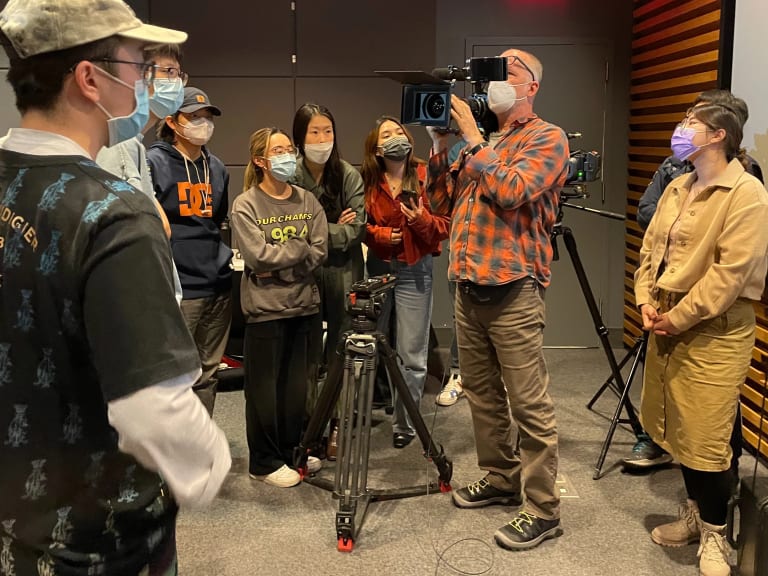 a man in a plaid shirt holding a large cinema camera is surrounded by his students who look on