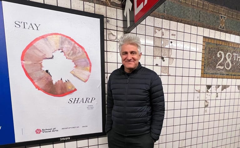 A man with gray hair, wearing a black jacket, smiling as he stands next to a poster in the subway.