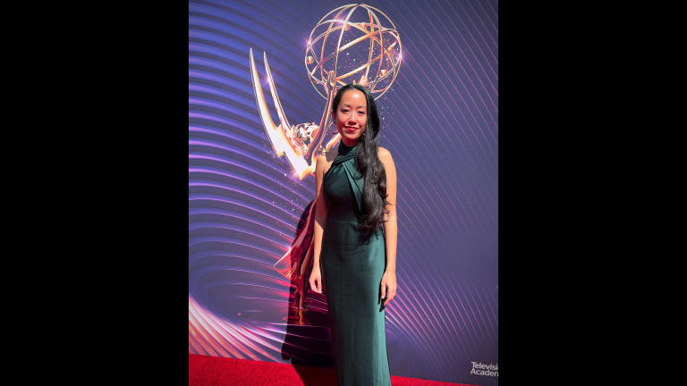 a woman in a green gown with long dark hair poses in front of a large photo of the Emmy award