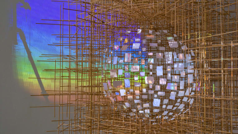 A sculpture of many small sticks arranged to create a sort of scaffolding where, in the center, is suspended a ball made of tiny white screens. On to each screen is projected a different image. A rainbow is projected onto the wall behind the work.