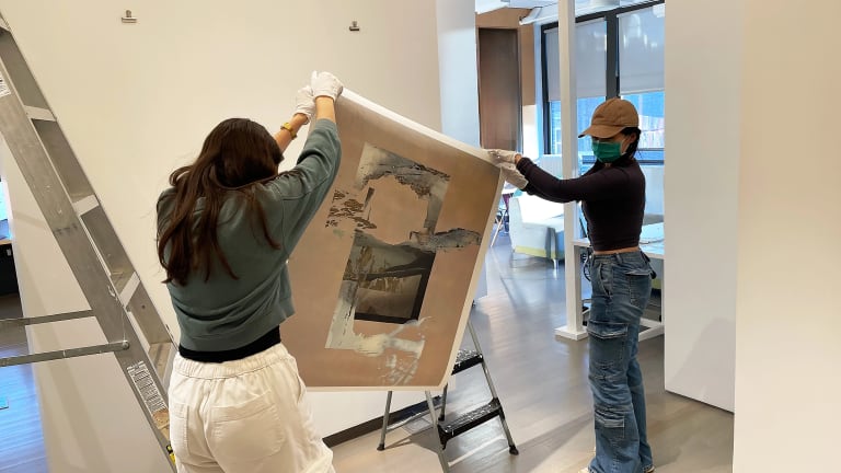 Two students hanging a work on paper on a wall in a gallery.