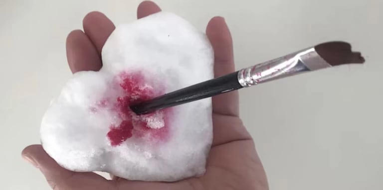 A hand holding a red-stained heart made of snow, with a paintbrush piercing its center