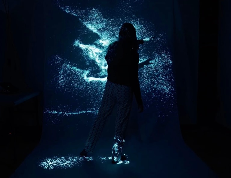 a person moves in front of a projection of abstracted blue particles in a dark studio space