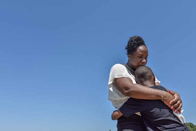 An African-American woman hugs her child. The background is a plain blue sky. She looks sad. We cannot see his face.