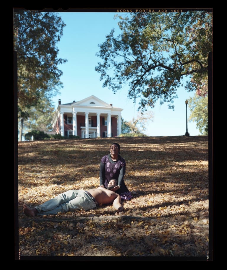 A color photograph of a black woman sitting in the grass cradling the head of a shirtless black man on her lap, as he lies flat in the grass. In the background is a brick building with white columns, trees, blue skies, and a streetlamp.