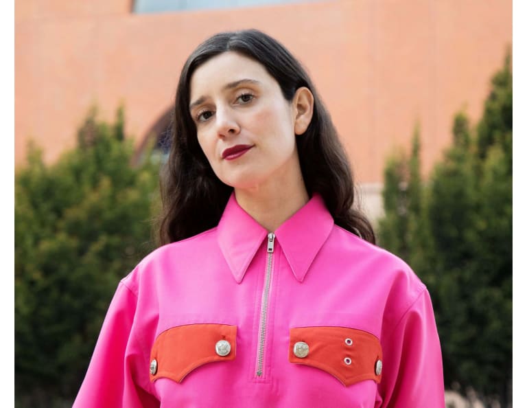 Image of a white woman in a pink zip up jacket with red lipstick and long brown hair looking at the camera with her head tilted 