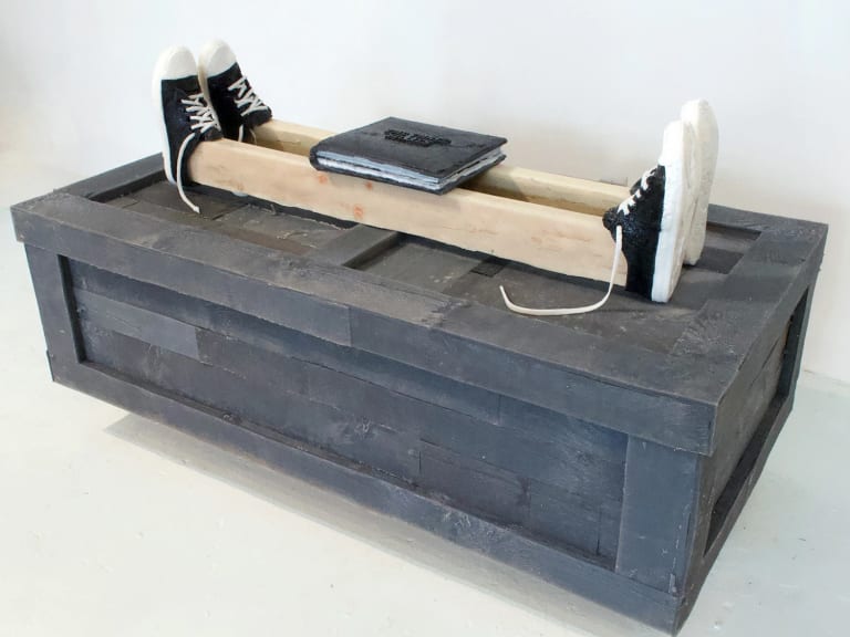 A silicone rubber sculpture of a photo album resting on two two-by-four planks, which in turn are resting on a gray shipping crate. The planks each wear two tennis shoes, one on each end, toes pointing upward