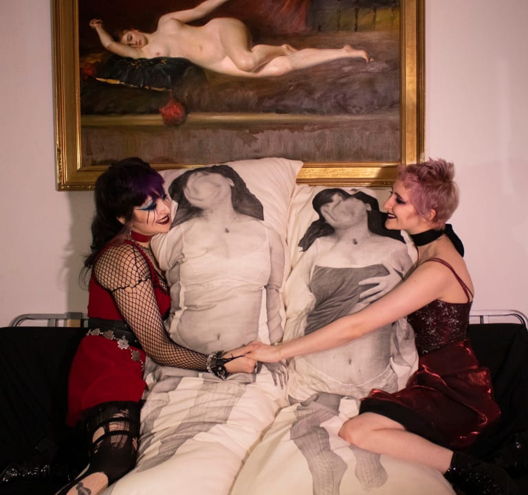 A painting of a reclining nude woman hangs on the wall, below which is a seat with two smiling women in goth clothing, facing each other and embracing pillows with life-sized black-and-white images of a woman in sleepwear
