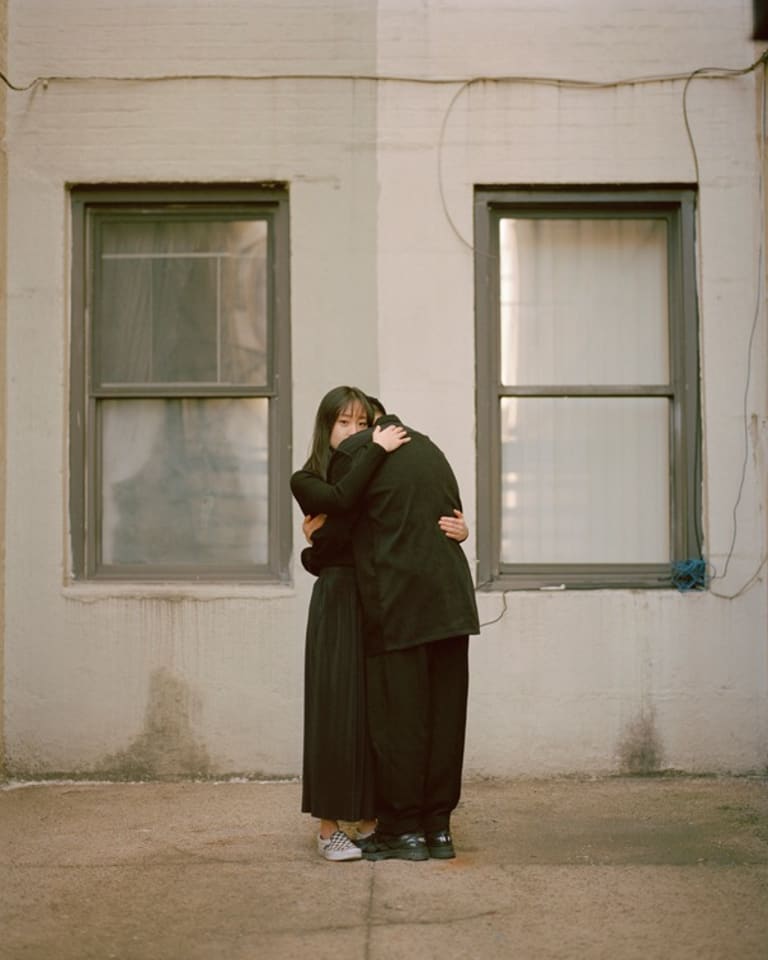 A color photograph of a young woman looking at the camera over the shoulder of a man she’s embracing. Both are dressed in all black. They are centered between two single windows on a building. The wall is painted two shades of grade and there is a line on the sidewalk, visually dividing the image, and the people in half.