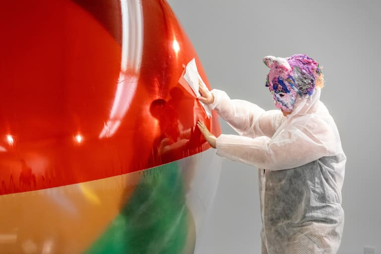A person wearing a full-body, semi-transparent protective suit with a colorful headpiece wipes a large inflated object in front of them