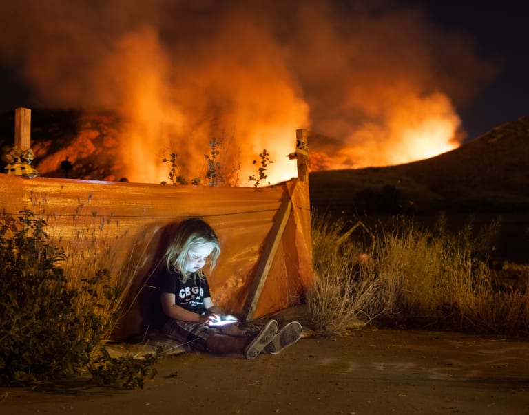 A photograph of a boy sitting outside and looking at a cellphone, as a wildfire burns in the background.