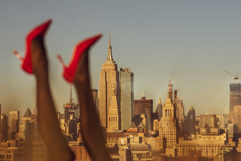 A color photograph. In the background, which is in focus, is a view of the New York City skyline with the Empire State building in the middle against a blue sky. In the foreground to the left, which is out of focus, are a pair of legs from the knees down, up in the air, wearing black tights and red high heel shoes.