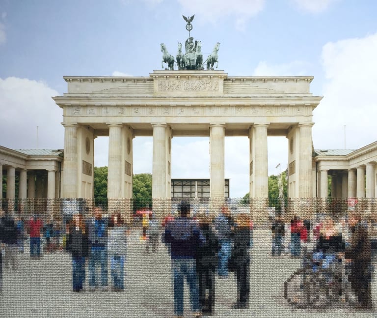 Color photograph of Brandenburg Gate in Berlin with people in front of it. The bottom third of the image, including all the people, is covered in embroidered cross-stitches in threads matching the colors of the image, giving it a pixelized look.