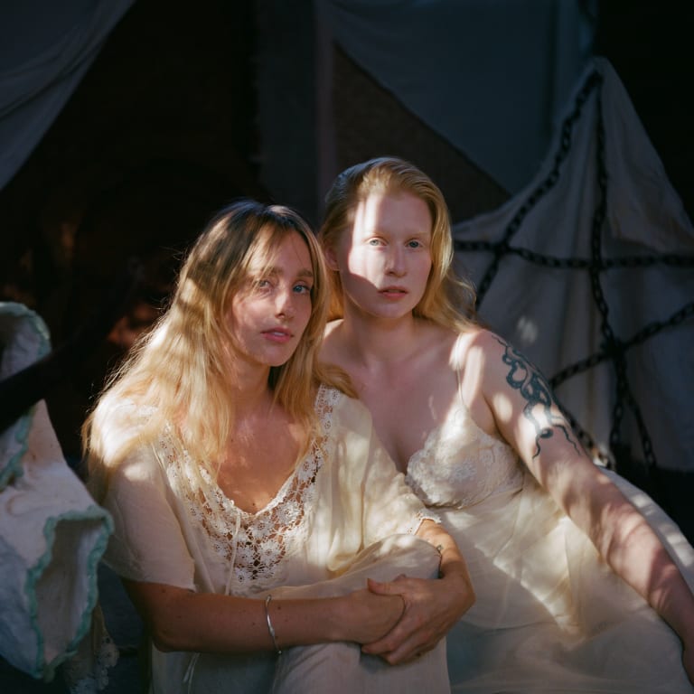 Color photograph of the artists in white dresses, sitting in a shaded area with scatters of light illuminating them from above and looking directly at the camera
