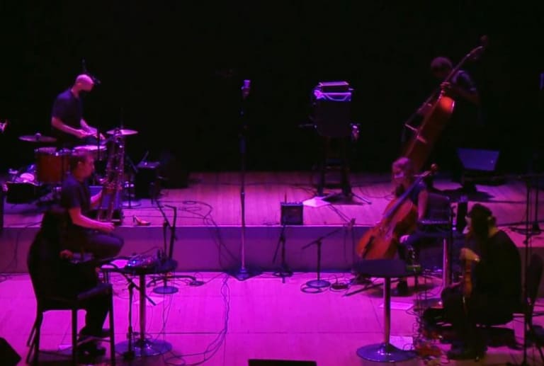 Six seated musicians playing both acoustic and electric instruments facing one another across a stage lit by purple spotlights in two rows of three performers each