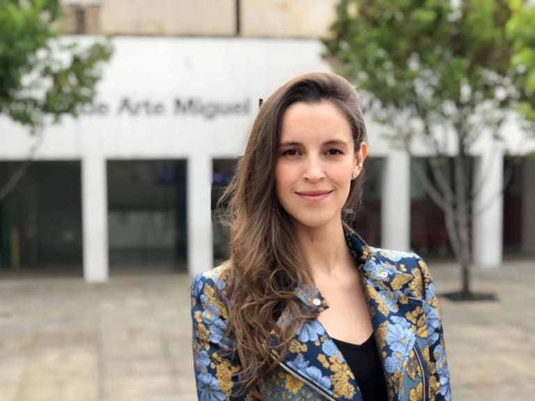 A woman stands in front of a white building wearing a blue and yellow floral blazer