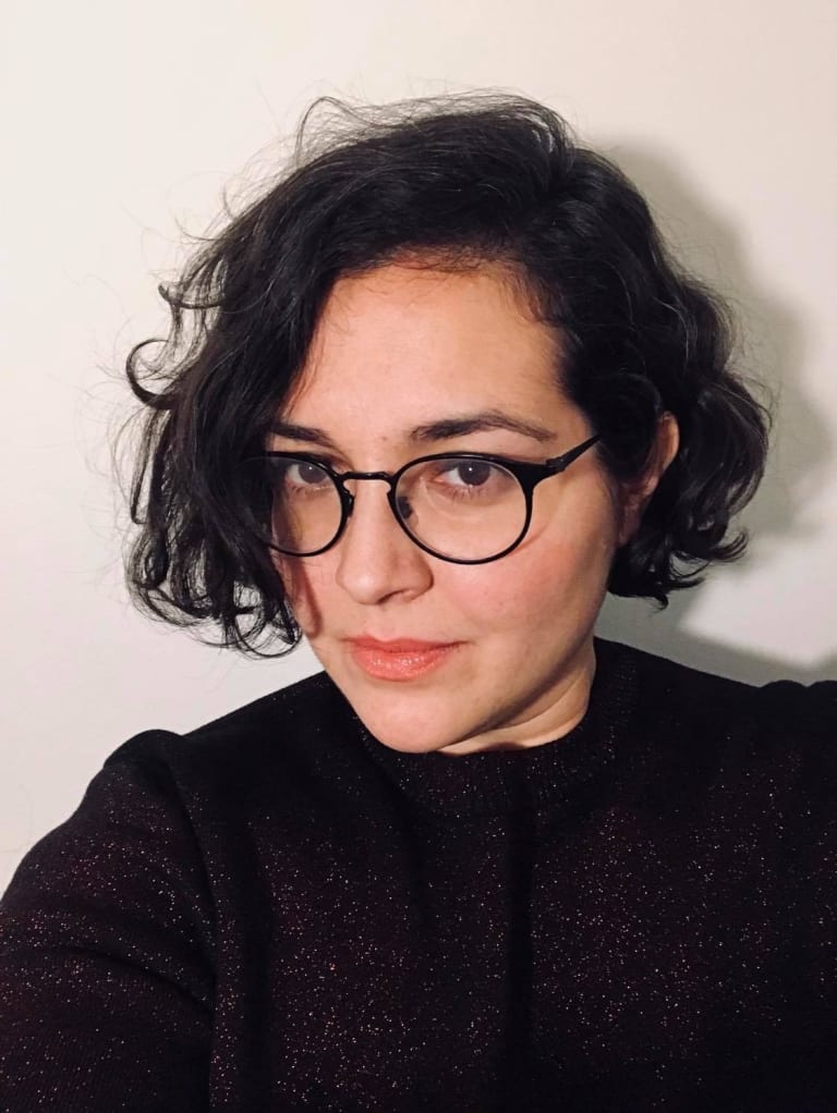 A woman with short, dark, curly hair wearing black framed glasses and a black shirt takes a selfie.