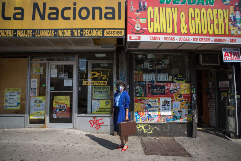 Image of a woman wearing a face mask, sunglasses, a blue coat, red shoes, and holding a briefcase standing in front of two stores with awnings that read "La Nacional" and "Candy & Grocery"