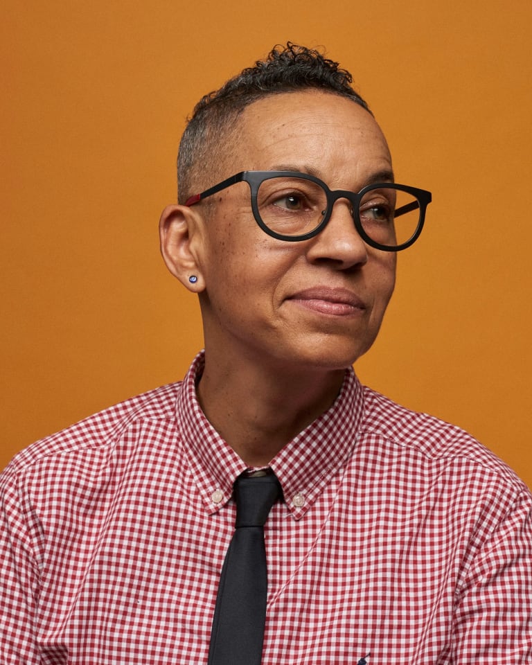 A studio portrait of photographer Lola Flash wearing a red and white plaid button up with a black tie.