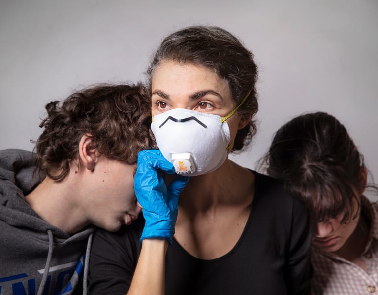 A photograph of a person in a face mask with two people hiding their faces in the person's shoulders