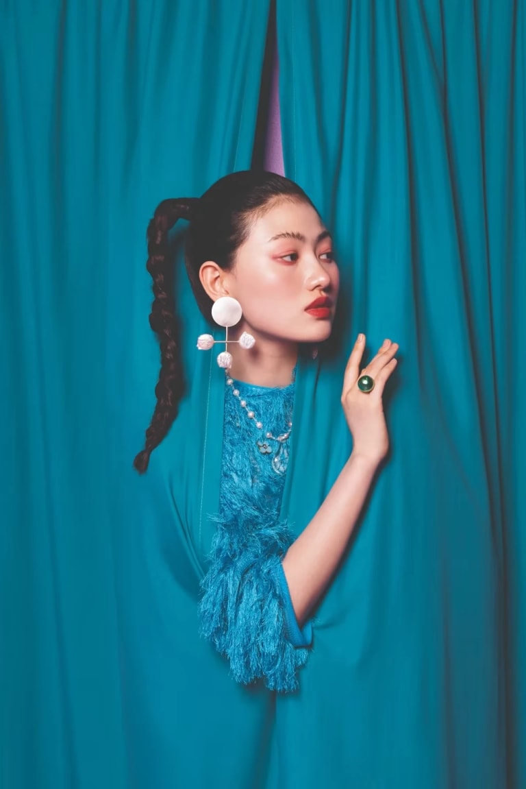A woman peers out from behind a set of blue curtains. She is wearing a large white earring and a round green ring